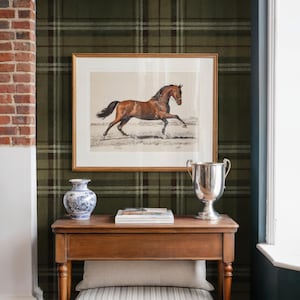May include: A framed print of a brown horse gallops above a wooden table. The table displays a silver trophy, a blue and white vase, and books. A striped cushion and a blue storage box are beneath. The room has a plaid wallpaper.