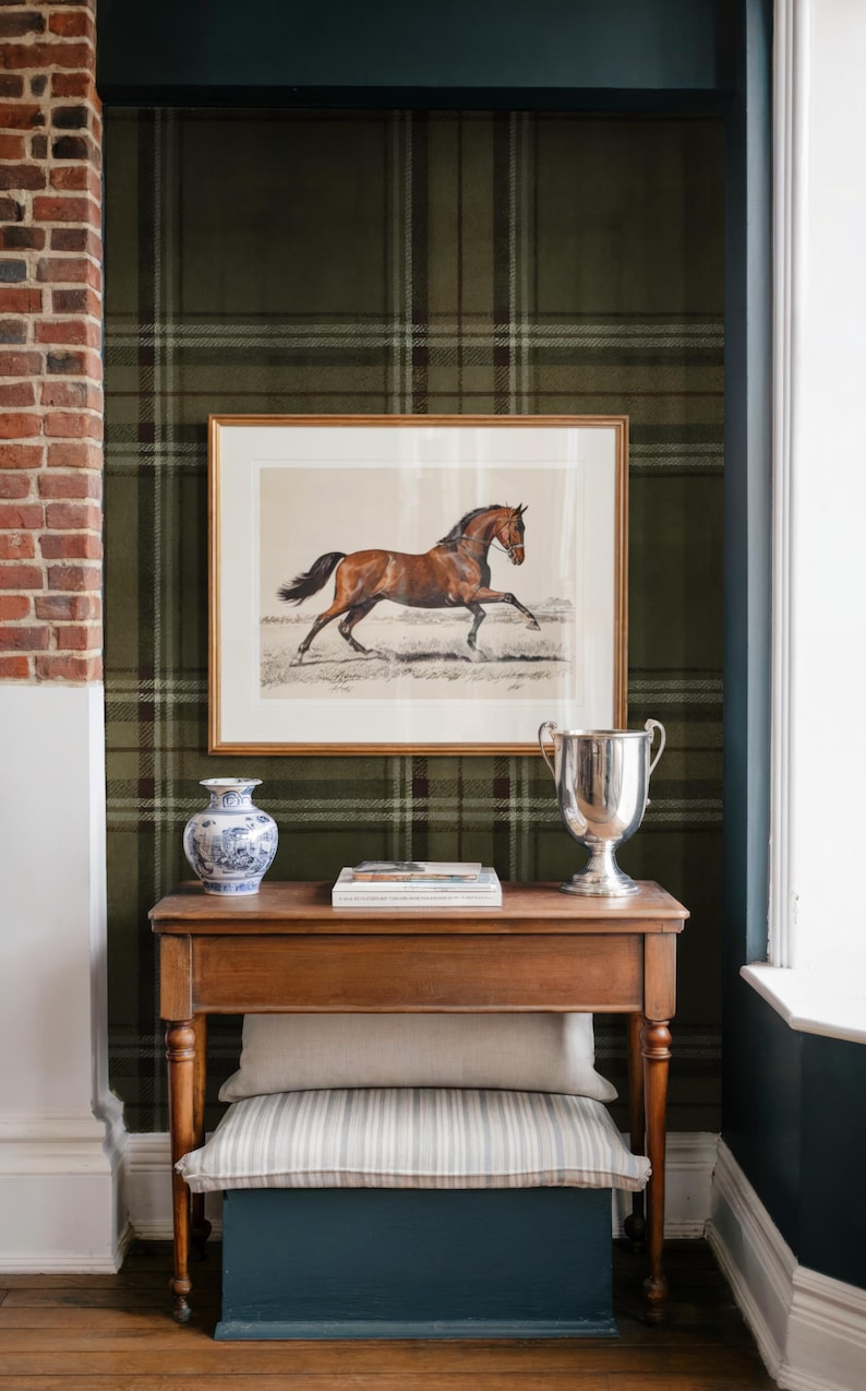 May include: A framed print of a brown horse gallops above a wooden table. The table displays a silver trophy, a blue and white vase, and books. A striped cushion and a blue storage box are beneath. The room has a plaid wallpaper.