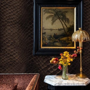 May include: An interior scene featuring a framed landscape painting with a black frame, a gold-toned lamp, and a small table with a marble top. A vase of flowers sits on the table. The wall has a textured, dark brown pattern.