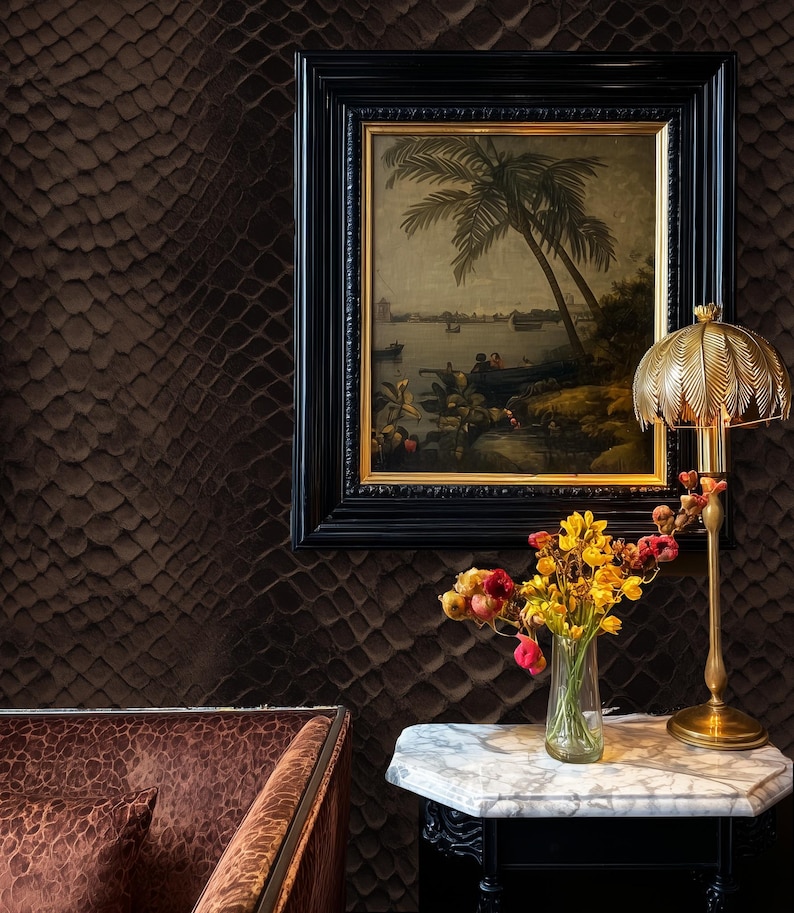 May include: An interior scene featuring a framed landscape painting with a black frame, a gold-toned lamp, and a small table with a marble top. A vase of flowers sits on the table. The wall has a textured, dark brown pattern.