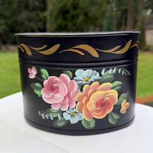 May include: A black, round metal container with a lid, featuring hand-painted roses in pink, yellow, and blue. Gold leaf accents adorn the top rim. The container is likely for storage or decorative purposes.
