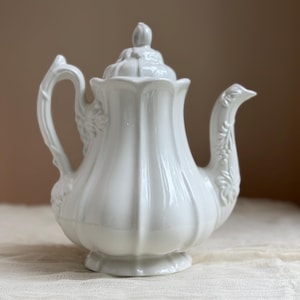 May include: A white ceramic teapot with a lid and a curved handle. The teapot has a fluted body and floral detailing on the handle and spout. The teapot is designed for serving tea.