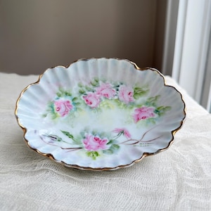 May include: Oval-shaped, hand-painted porcelain dish with a scalloped edge and gold trim. The dish features a delicate floral design of pink roses and green leaves on a pale blue and white background. A charming decorative piece.