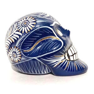 May include: A ceramic skull figurine in a deep blue glaze, decorated with intricate hand-painted white floral patterns and delicate gold glitter accents. The skull features a smooth, glossy finish with visible teeth and eye sockets, viewed from a side profile.