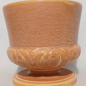 May include: A peach-colored ceramic planter with a textured, matte upper body and a glossy, raised scrollwork pattern around the base. The vessel features a wide, open top and sits on a circular pedestal foot, showcasing a consistent warm tone.