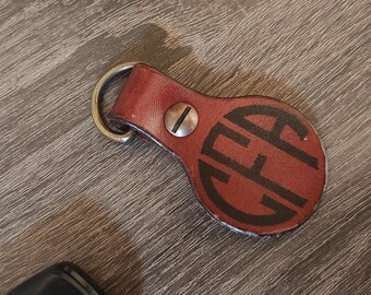 Op de afbeelding: Een bruine leren sleutelhanger met een messing ring en een rond, gemonogrammeerd label met de letters "CEA" in zwart.