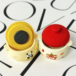 May include: Two miniature ceramic pots with lids. One pot has a yellow lid and a panda illustration, the other has a red lid and a cartoon animal illustration. Both pots are cream-colored with small handles.