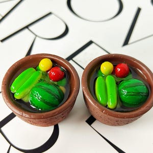 May include: Two miniature brown bowls filled with a clear liquid and colorful miniature food items. Each bowl contains a green watermelon, two green pickles, a yellow ball, and a red tomato. The bowls are set against a white background.