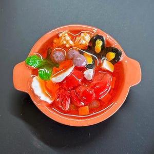 May include: A small ceramic bowl filled with a colourful dish. The contents include red tomatoes, orange carrots, white and purple spheres, and green leafy elements, all in a clear broth. The bowl is a warm orange colour.