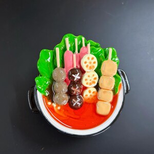 May include: A colourful, miniature food sculpture of a Korean hot pot. The dish features skewers of meatballs, mushrooms, lotus root, and tofu, along with lettuce and a red broth. The pot has a black rim and handles.
