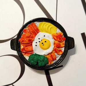 May include: A miniature replica of a Korean bibimbap dish. The dish includes a fried egg, rice, vegetables, and meat, presented in a black bowl. The food items are brightly coloured, with a focus on the yellow egg yolk and the red meat.