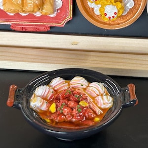 May include: A miniature black ceramic bowl filled with a vibrant red stew, topped with white and pink dumplings. The bowl has brown handles and sits in front of other decorative food items.