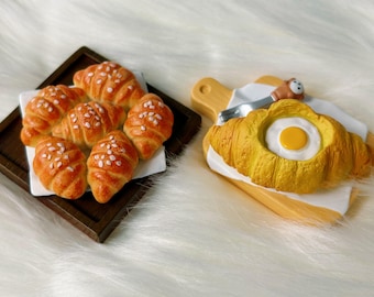 May include: An image of miniature croissants, some with white sprinkles, arranged on a small wooden tray. Another croissant is on a wooden cutting board with a fried egg inside. A knife and a small bear are also on the cutting board.