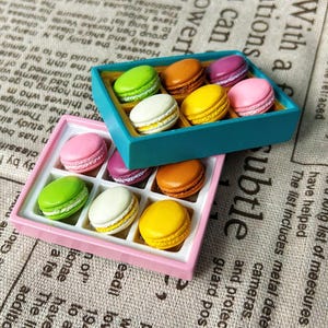 May include: Two boxes of colourful macarons. One box is teal, the other is pink. Each box contains nine macarons in various colours, including green, orange, purple, yellow, white, and pink. The macarons are arranged in a grid pattern.