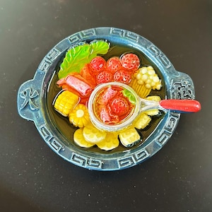 May include: A blue decorative plate with a detailed miniature food arrangement. The plate features a variety of colorful food items, including corn, tomatoes, and a small bowl of soup. The plate has a decorative border and handles.