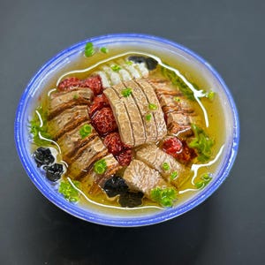 May include: A bowl of soup with a blue rim, filled with broth, sliced meat, red berries, and green herbs. The soup has a yellow-orange hue, and the ingredients are arranged in a visually appealing manner. The bowl is set against a dark background.