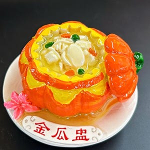 May include: A ceramic dish in the shape of a pumpkin, filled with yellow soup and white ingredients. The dish is orange and yellow, with a white plate base and a pink flower accent. The dish has Chinese characters.