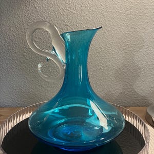 May include: A turquoise glass jug with a clear, looped handle. The jug has a wide base and a flared spout. It sits on a silver-rimmed tray with a black interior. The background is a neutral grey.
