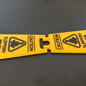 May include: A yellow caution sign with black text that reads "Caution Slippery When Reading". The sign is split in half and is made of a flexible material.
