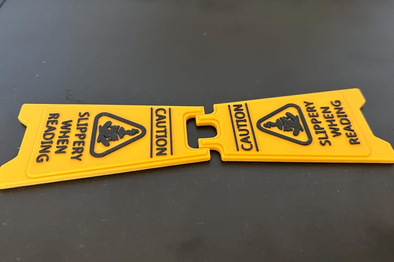 May include: A yellow caution sign with black text that reads "Caution Slippery When Reading". The sign is split in half and is made of a flexible material.