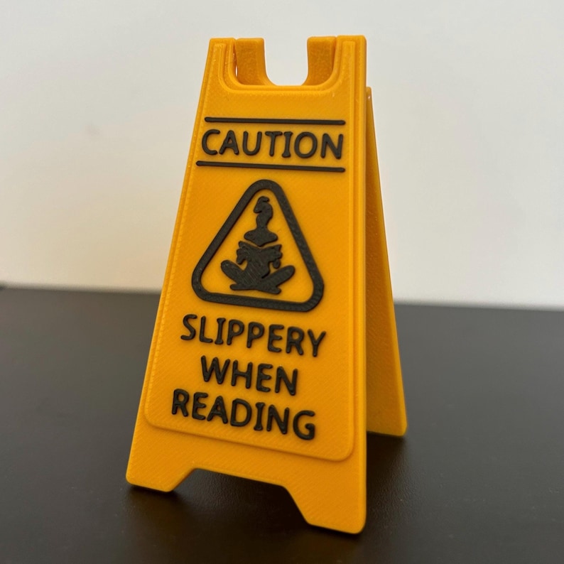 May include: A yellow caution sign with a black silhouette of a person reading a book. The sign reads "Caution Slippery When Reading".