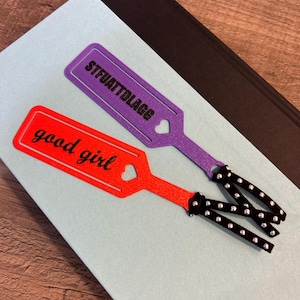 Peut inclure: Deux marque-pages violet et rouge avec une d&eacute;coupe en forme de c&oelig;ur et un ruban noir &agrave; pois argent&eacute;s. Le marque-page violet dit "STFUATDLAGG" et le marque-page rouge dit "good girl".