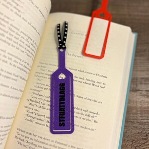 Peut inclure: Signet de livre violet avec des paillettes et le texte "STFUATTDLAGG" en lettres noires. Le signet a la forme d'un long rectangle avec un c&oelig;ur d&eacute;coup&eacute; en haut.