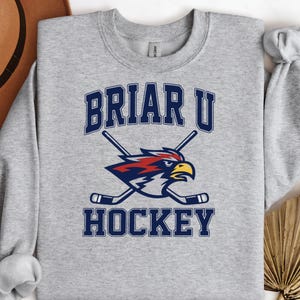 May include: A heather gray crewneck sweatshirt featuring a dark blue graphic that reads BRIAR U HOCKEY in a collegiate font. The design includes a stylized bird head in red, white, and blue, positioned behind two crossed hockey sticks in blue.