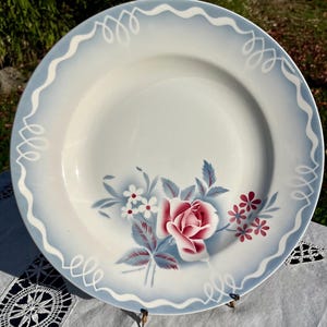 Puede incluir: Un plato de cerámica blanca vintage con un borde festoneado azul claro y un diseño floral. El centro presenta una rosa rosa con hojas azul-gris y pequeñas flores blancas y rojas. Vajilla decorativa.