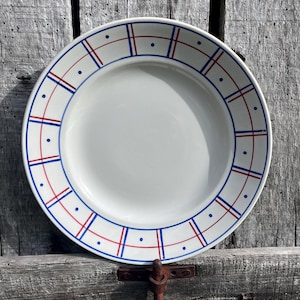 Könnte beinhalten: Ein weißer Keramikteller mit einem blau-roten Gittermuster am Rand. Der Teller hat ein schlichtes, klares Design mit einem leicht erhöhten Rand. Der Teller steht vor einem verwitterten Holzgrund.