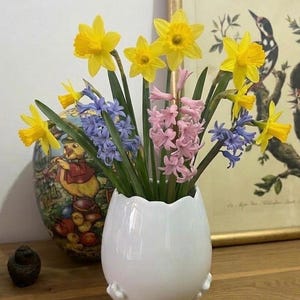Pode incluir: Um vaso de cerâmica branco em forma de ovo com um buquê de narcisos amarelos, jacintos cor-de-rosa e jacintos azuis. O vaso tem três pés pequenos. Uma impressão emoldurada e um vaso decorativo em forma de ovo estão no fundo.
