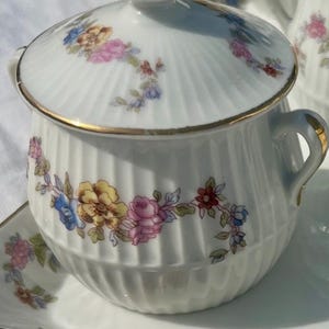 Puede incluir: Azucarero de porcelana blanca con tapa, ribete dorado y estampado floral en rosa, amarillo y azul. El cuenco tiene un diseño acanalado y asas pequeñas. Parte de un juego de té.