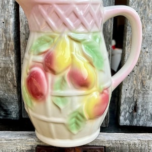 Pode incluir: Jarro de cerâmica vintage com alça e borda rosa claro. O jarro é decorado com frutas e folhas em relevo em tons de vermelho, amarelo e verde. A parte superior do jarro tem um design de tecido de cesta.