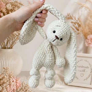 May include: A handmade, light green crochet bunny toy with long floppy ears, a small brown nose, and black eyes. The bunny is soft and plush, with a textured surface. The toy is held up by a hand.