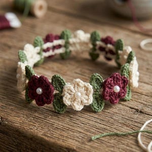 May include: A handcrafted crochet flower crown featuring cream, burgundy, and green flowers with pearl centers. The floral headband is arranged on a wooden surface, showcasing the intricate details of the handmade accessory.