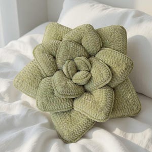 Könnte beinhalten: Ein hellgrünes, plüschiges Zierkissen in Form einer Sukkulentenpflanze. Das Kissen hat ein strukturiertes, samtiges Aussehen und liegt auf einem weißen Bett. Das Design ahmt die geschichteten Blütenblätter einer Sukkulente nach.