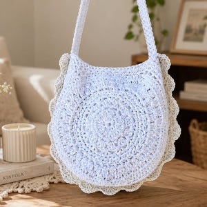 May include: A white crochet bag with a circular pattern and a scalloped edge. The bag has a long strap. The bag is displayed on a wooden table. The bag is part of a home decor theme.