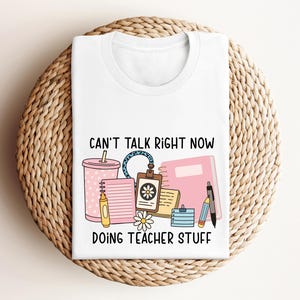 May include: White t-shirt with the text "CAN'T TALK RIGHT NOW DOING TEACHER STUFF" and a graphic of a pink tumbler, notebook, pen, and other school supplies. The shirt is displayed on a woven, circular placemat.