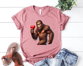 May include: A dusty rose t-shirt featuring a graphic of a brown Bigfoot holding a red heart. The shirt is paired with blue jeans and brown sandals. A small green plant is visible in the background. The shirt is a casual wear item.