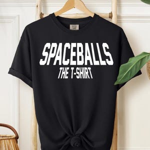 May include: Black t-shirt with the text "SPACEBALLS THE T-SHIRT" in white, displayed in a bold, blocky font. The shirt is hanging on a wooden rack, with a knot tied at the bottom.