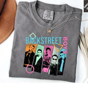 May include: A gray Comfort Colors t-shirt featuring the text "BACKSTREET BOYS" in teal and pink, with images of the band members. A silver necklace with a pendant is draped on the shirt.