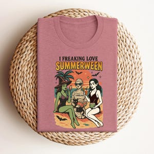 May include: A dusty rose t-shirt with the words "I Freaking Love Summerween" above a graphic of three figures on a beach. The figures include a witch, a mummy, and a woman in a swimsuit, with bats and palm trees.