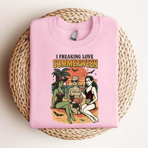 May include: Pink sweatshirt with the text "I Freaking Love Summerween" and a graphic of three women dressed as a witch, mummy, and vampire on a beach. The graphic includes palm trees, bats, and a black cat.
