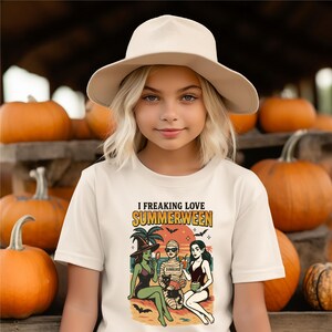May include: A young person wearing a cream-colored t-shirt with the text "I Freaking Love Summerween" and a graphic of three cartoon figures on a beach. The person is also wearing a beige hat, with a background of pumpkins.