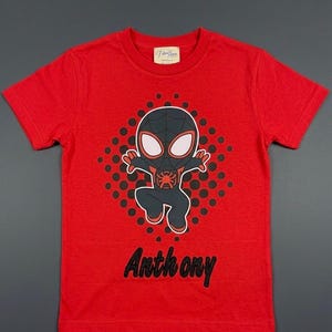 May include: A red t-shirt featuring a cartoon illustration of a superhero character. The character is black and red with white eyes, set against a black dotted background. The name "Anthony" is printed in black script below the graphic.