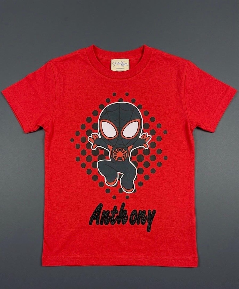 May include: A red t-shirt featuring a cartoon illustration of a superhero character. The character is black and red with white eyes, set against a black dotted background. The name "Anthony" is printed in black script below the graphic.