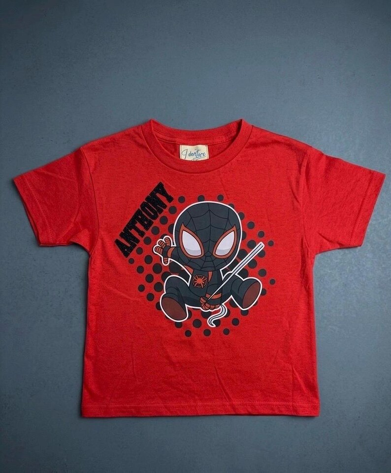 May include: Red t-shirt featuring a cartoon Spider-Man character with the name "ANTHONY" printed on the left side. The graphic includes a black and gray Spider-Man design with a web. The shirt is made of a soft material.