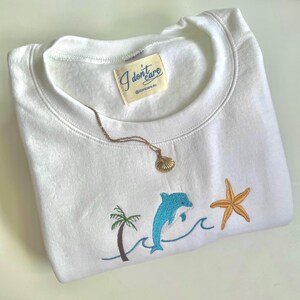 Puede incluir: Sudadera blanca con diseño bordado de temática oceánica, que incluye un delfín azul, una palmera y una estrella de mar. Un collar con colgante de concha dorada descansa sobre la sudadera. La etiqueta dice "I don't care".