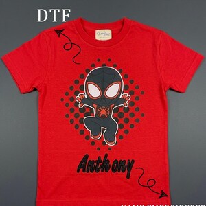 May include: Red t-shirt featuring a black and red cartoon character design with the name "Anthony" in black glitter lettering. The shirt also has the text "NAME EMBROIDERED ON GLITTER".