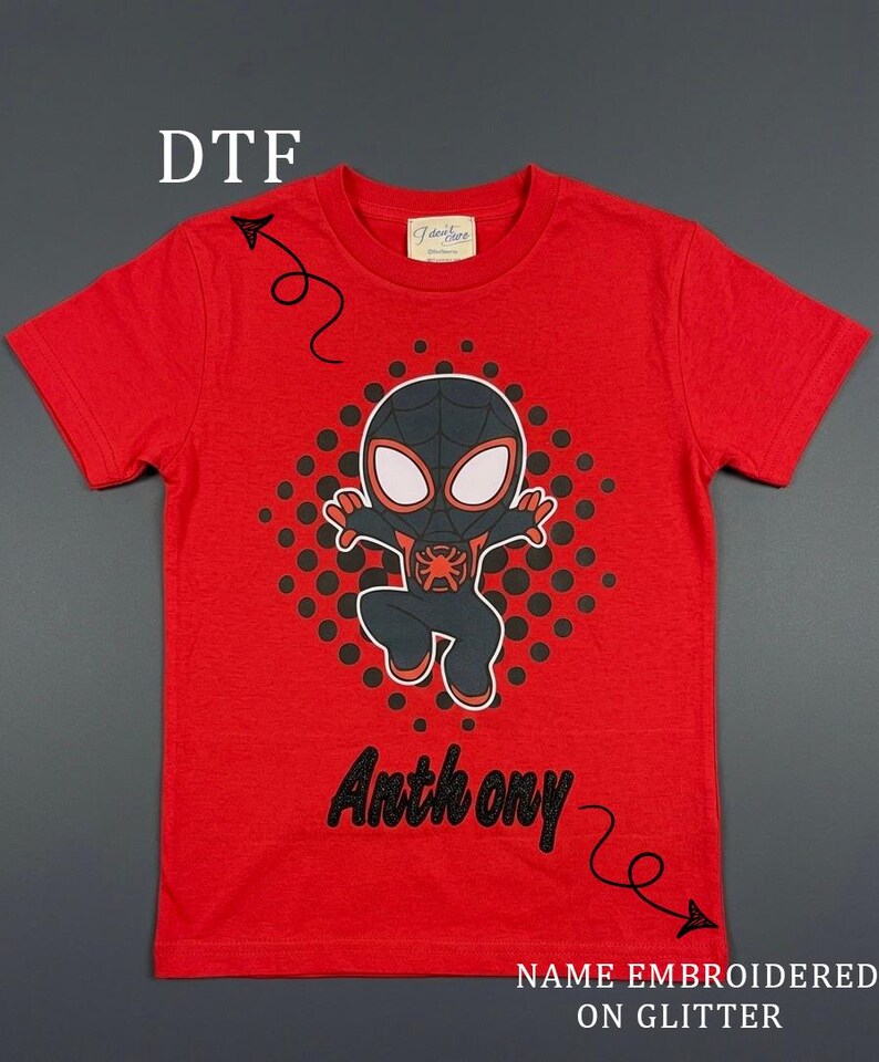 May include: Red t-shirt featuring a black and red cartoon character design with the name "Anthony" in black glitter lettering. The shirt also has the text "NAME EMBROIDERED ON GLITTER".
